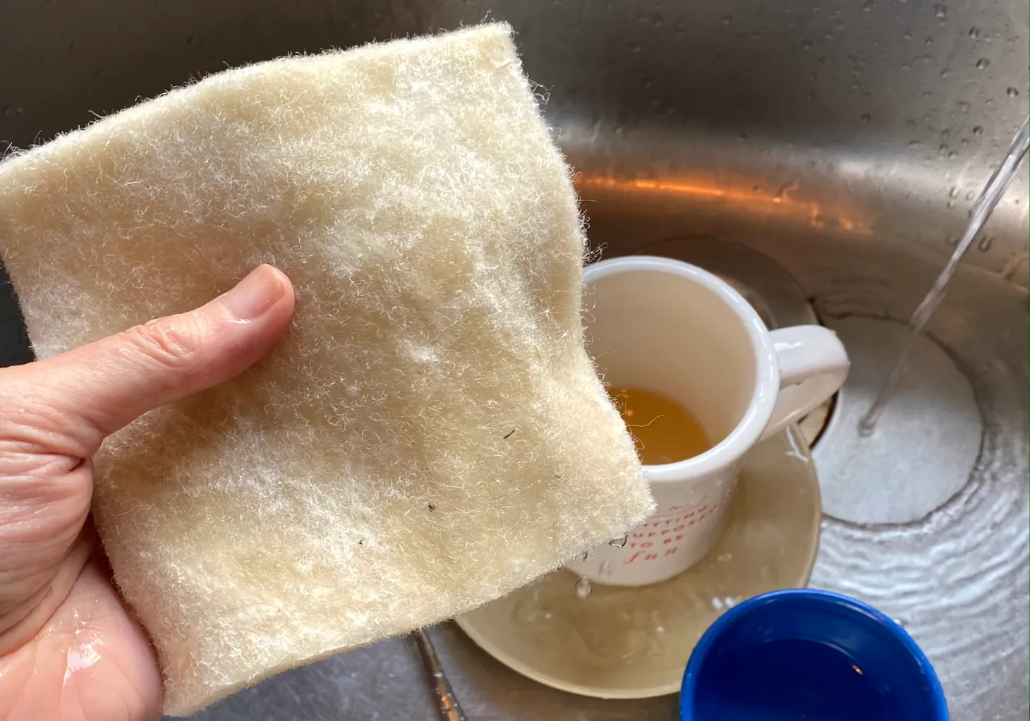 A hand holds a wet square of thick wool felt over a sink of dirty dishes.
